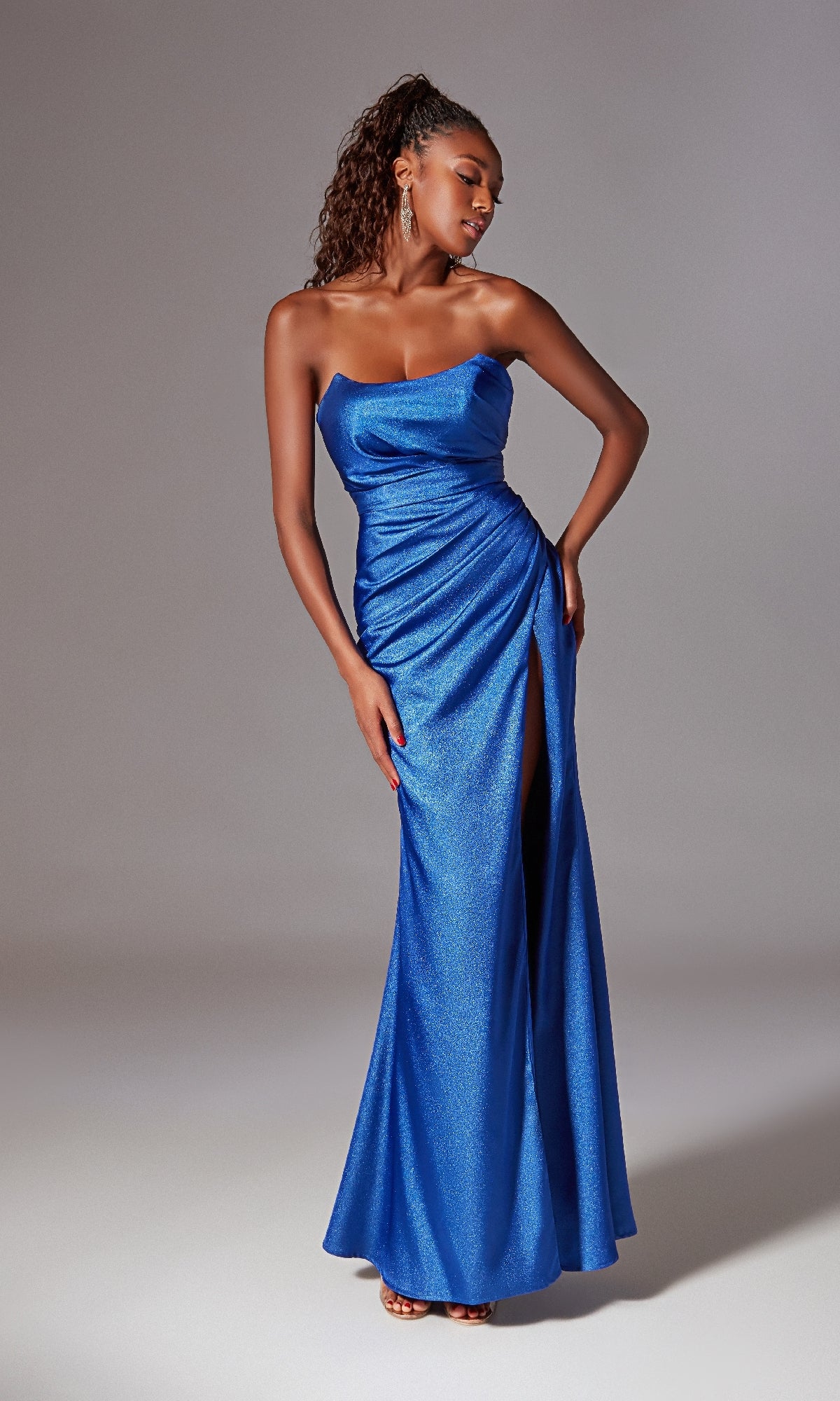 A woman models the Laced Strapless Long Prom Dress: Aerie Couture 247, a metallic blue gown with a thigh-high slit, posing against a plain light gray background.
